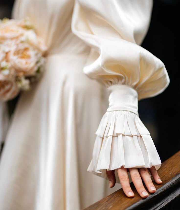 closeup of a bride in an elegant, ivory satin wedding dress with puffy sleeves and frilled cuffs, holding a bouquet of light orange roses, her hand resting on a wooden surface. the dress has soft folds and a luxurious texture, exuding a vintage, romantic, and soft aesthetic.