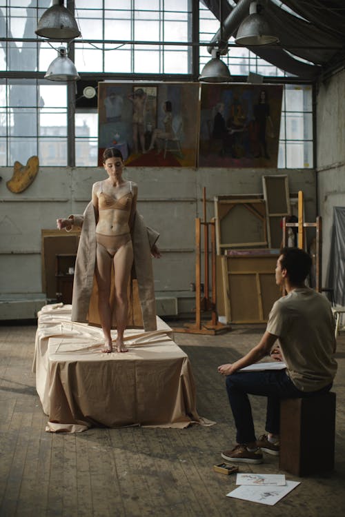 the image shows a female model standing on a draped surface in a spacious art studio.  she is wearing a light beige bra and panties, with a long, matching robe draped over her shoulders.  a male artist sits on a wooden stool in the foreground, observing her and seemingly sketching. the studio has a rusticindustrial feel with large windows, exposed beams, and art supplies and canvases scattered about. the overall atmosphere is calm and focused, suggesting a moment of artistic creation. the color palette is muted, predominantly beige and neutral tones, emphasizing the intimacy of the scene.