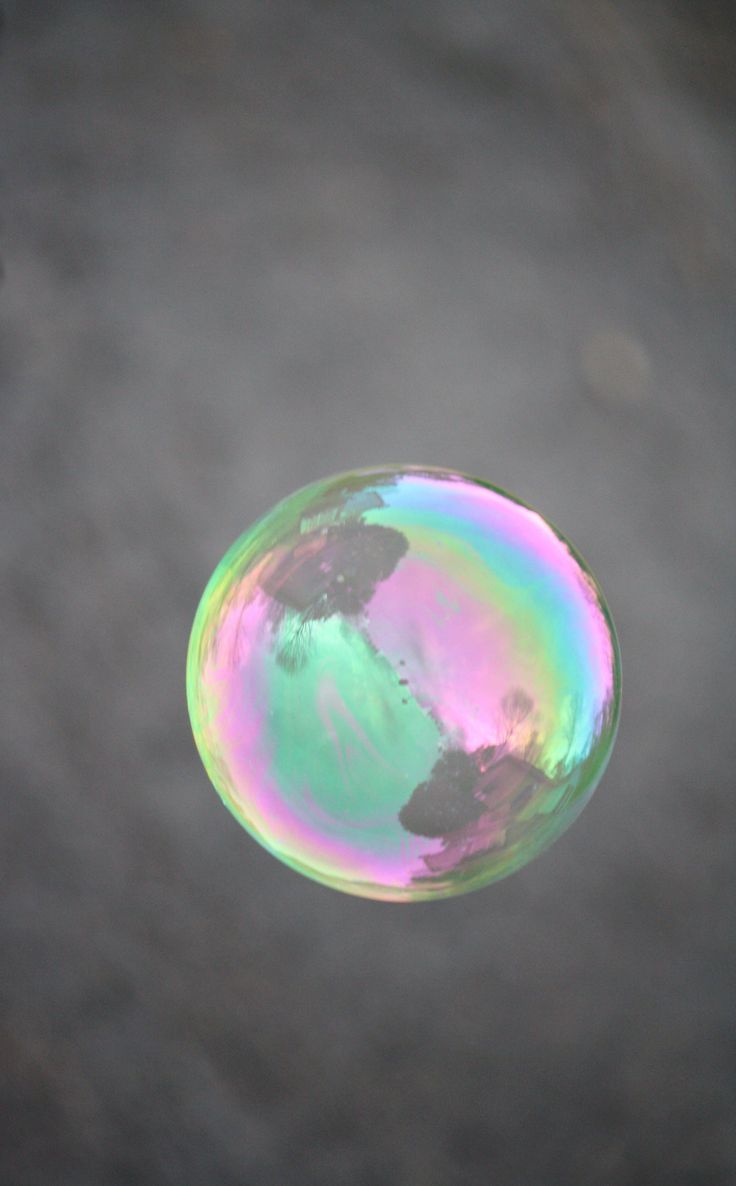 a colorful iridescent soap bubble floats in the air against a blurry gray background. the bubble is round and shiny, and it reflects the light.