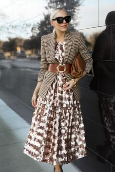 a blonde woman wearing black sunglasses, a checkered blazer and a sequined dress, poses outside on a city street. she has a brown purse in one hand, and she is wearing a brown leather belt with a golden buckle. she is looking at the camera and smiling. the photo is taken during the day.