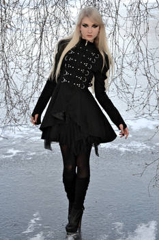 young blonde woman in a black gothicstyle dress and boots poses outdoors in a snowy winter landscape.  the dress has a layered skirt and decorative buckles. the scene is somewhat dramatic and mysterious due to the dark clothing and wintry setting. the woman's pale complexion contrasts with the black dress.  the background features bare, snowcovered trees.