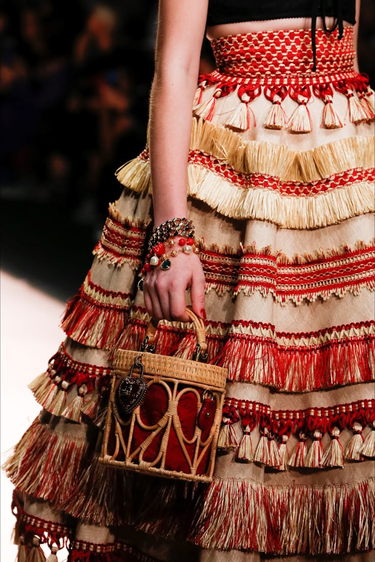a woman is captured on a runway, showcasing a tiered skirt with red and beige tassels, paired with a wicker bag. her wrist is adorned with multiple bracelets.