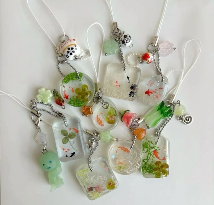 a collection of handmade resin charms and keychains featuring miniature goldfish and plant life inside clear resin. the charms have various shapes and are attached to mobile phone straps. some charms have additional beads or figurines attached. the style is cute and kawaii.
