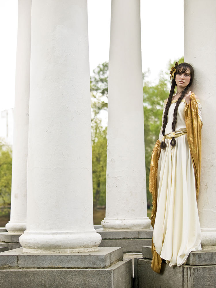 a young woman in a long, flowing offwhite gown with gold accents stands between white pillars. she has long brown hair in braids and a serene expression.  the setting is outdoors in a parklike area with trees visible in the background. the image has a classical or historical feel.