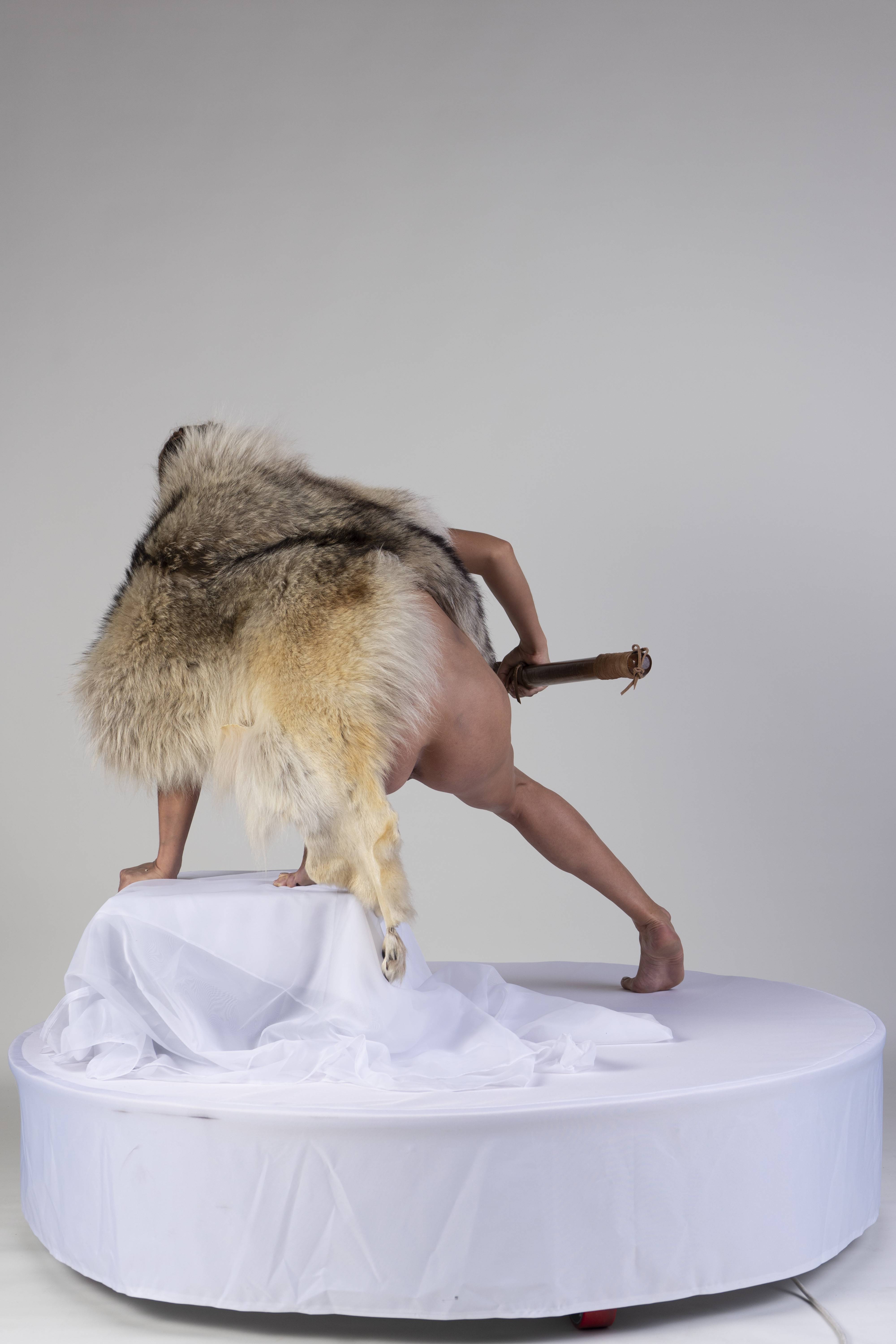 an artistic image features a nude person, partially covered by a fur garment, positioned on a round white table covered with a white cloth. the person is leaning forward with one hand and one foot on the table, while the other leg is extended back and holds a wooden object. the background is a plain white surface.