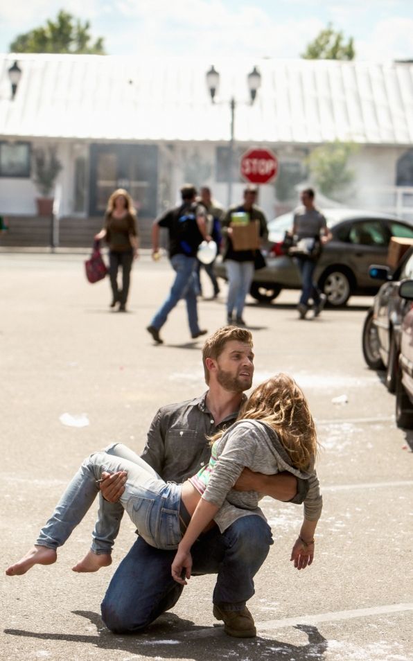 a man is holding a woman, who appears to be injured, in his arms. the man is helping her in a city street. there is a commotion around them with other people running in the background.