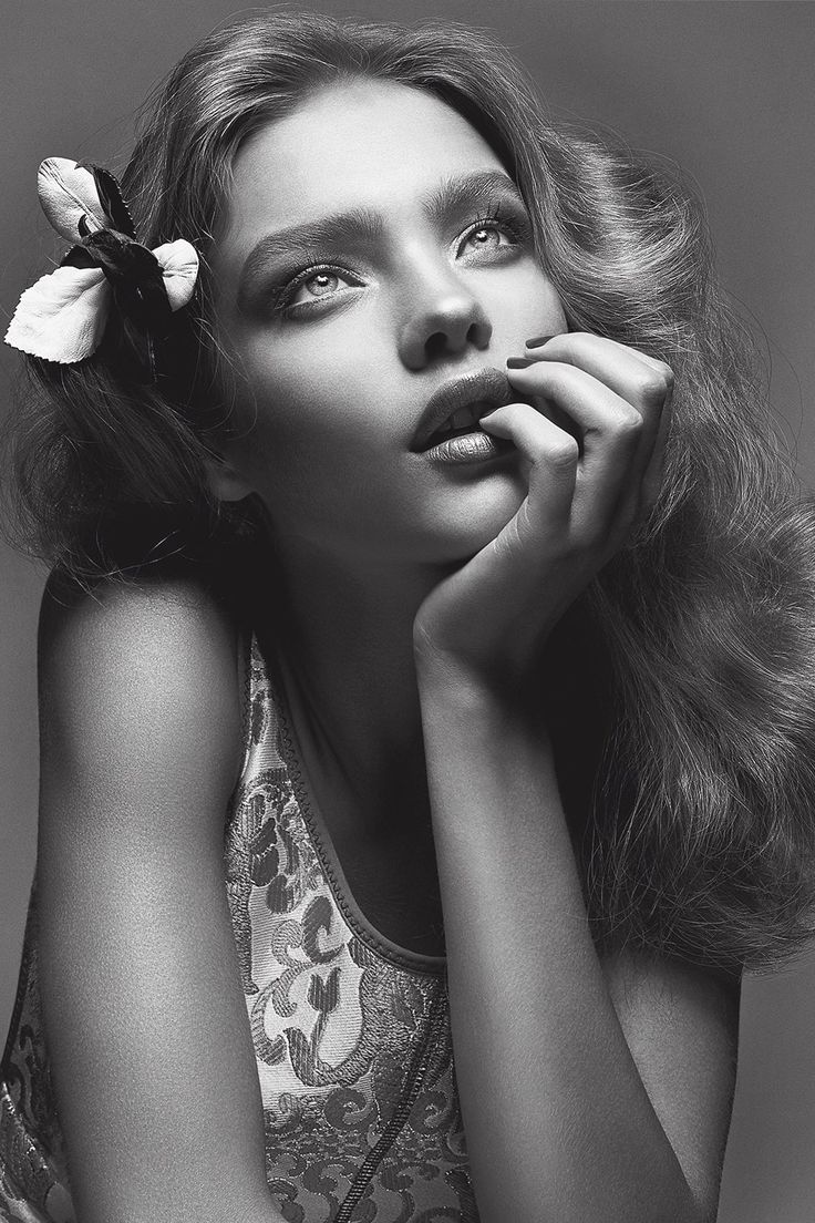 a black and white portrait of a beautiful young woman with long wavy hair, wearing a sleeveless dress with a floral pattern. the woman is gazing upwards with a thoughtful and wistful expression. she is holding her hand to her chin, and a flower is tucked behind her ear. the lighting is soft and diffused, creating a dreamy atmosphere.