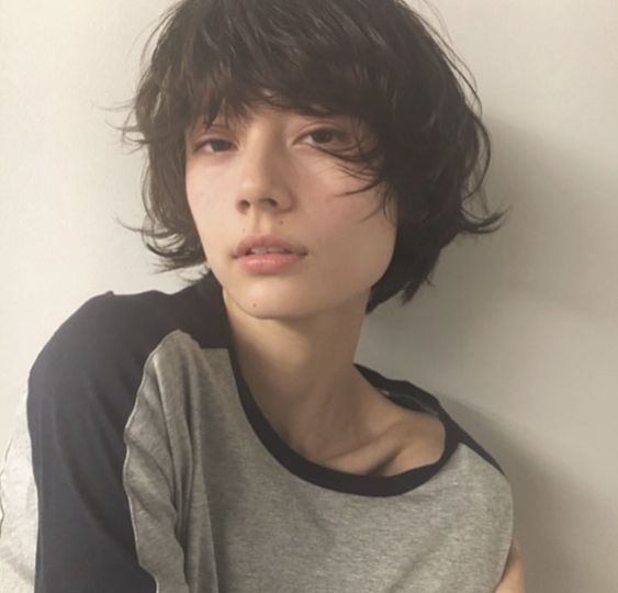 the image shows a closeup portrait of a young woman with short, tousled dark hair. she has a neutral expression on her face and is wearing a simple gray and black tshirt. the background is a plain white wall. the lighting is soft and natural.
