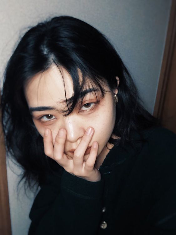 a young asian woman with black hair, wearing a black top, covers her mouth with her hand while looking at the camera. the lighting is dim and the environment appears to be indoors.