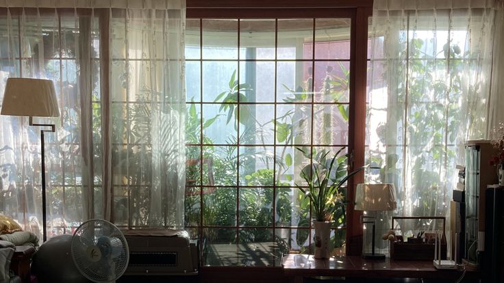 the image depicts a sunlit room with a large window dressed in sheer, lacelike curtains.  the window looks out onto a variety of lush green plants, creating a tranquil, gardenlike view from indoors.  inside the room, there's a floor lamp, a table lamp, a desk with what appears to be a small bookshelf or storage unit, a desk fan, and a what looks like a small portable air conditioner. the overall atmosphere is peaceful, serene, and cozy, suggesting a calm and comfortable home interior. the sunlight streams through the curtains, casting shadows and highlighting the textures and details of the room.