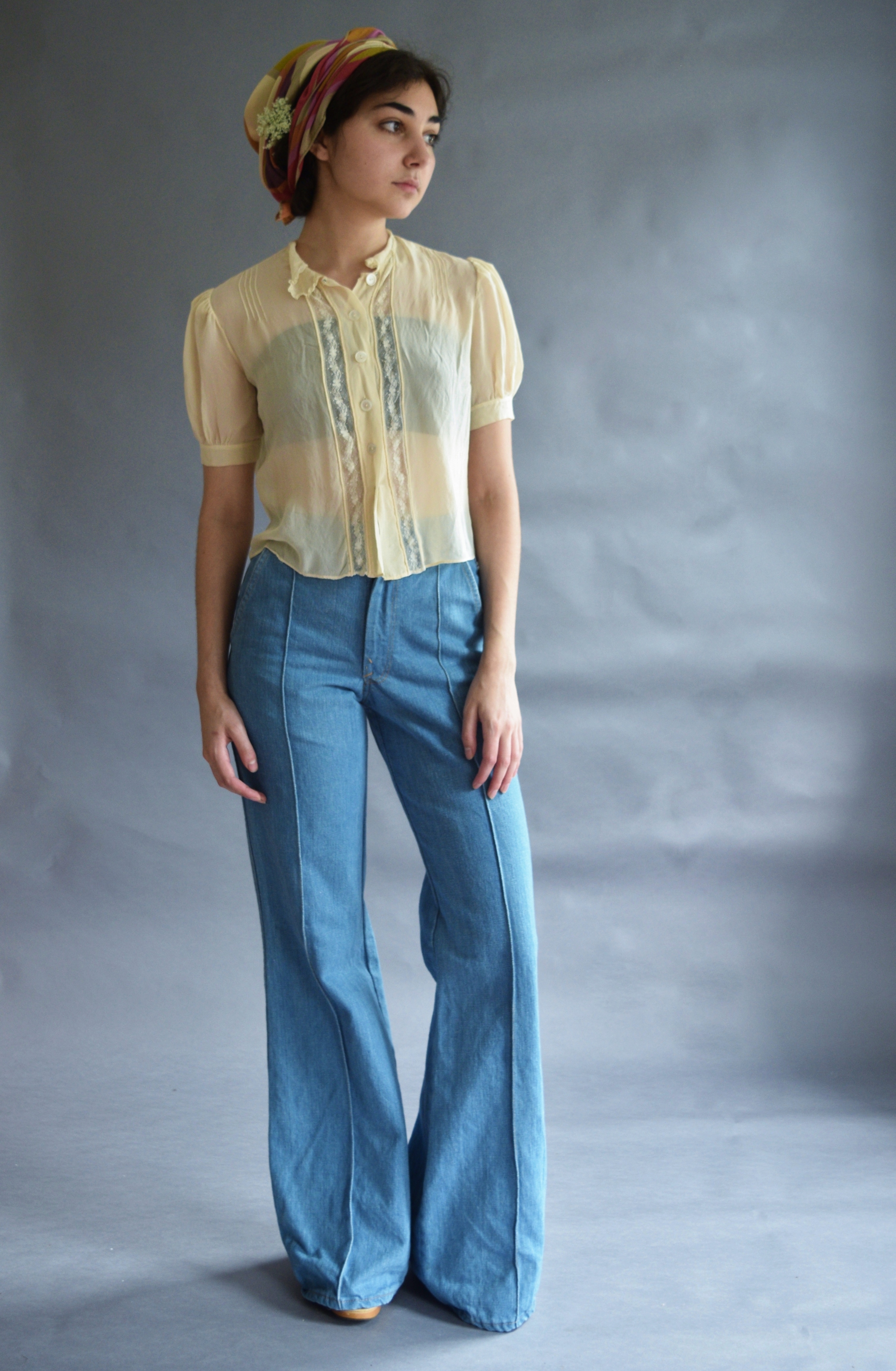 a full shot of a young woman modeling a vintage 1970s outfit. she's wearing a light yellow sheer blouse with delicate lace detailing, paired with widelegged, light blue bellbottom jeans.  her hair is tied up with a colorful patterned headscarf. the background is a muted gray studio backdrop. the overall style is reminiscent of 1970s fashion.