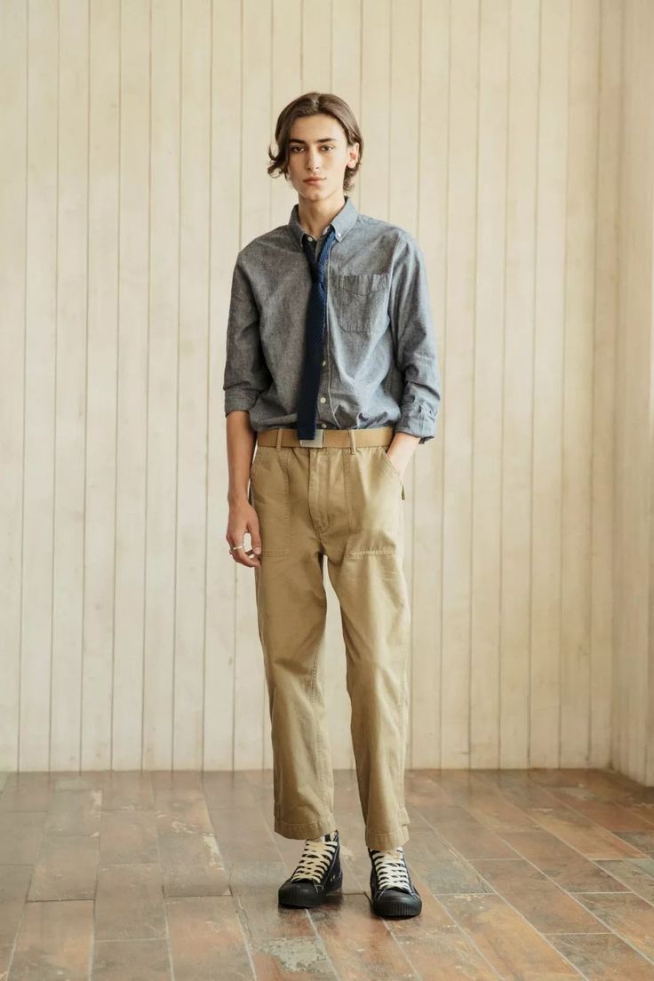 a young man stands in front of a light wood paneled wall. he is wearing a gray chambray shirt, a dark tie, beige work pants, and black hightop sneakers. he is looking directly at the camera. the image is a full body shot and is taken in a studio setting.