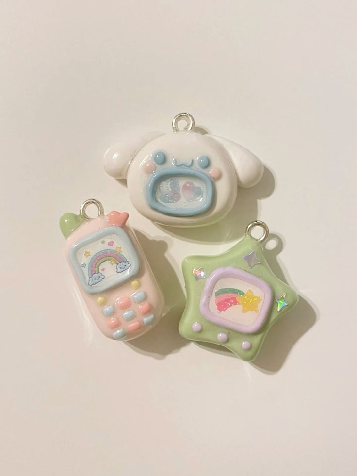 the image shows three kawaiistyle charms. from top to bottom, they include a cinnamorollshaped charm with a blue frame around its face containing glitter, a pink cell phone charm with a rainbow on the screen and buttons, and a green starshaped charm with a framed picture containing a shooting star. all charms are pastelcolored and have a metal ring at the top for attachment.