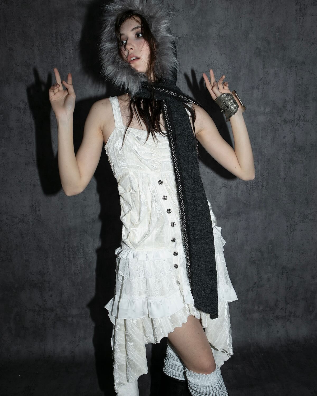 a young asian woman model poses in a studio against a dark textured wall. she is wearing a white ruffled dress with a grey furtrimmed hood and scarf. the scarf is long and drapes down to her knees. she is also wearing a bracelet on her right wrist. she is making a peace sign with both hands. the overall style is elegant and pretty, with a hint of winter fashion.