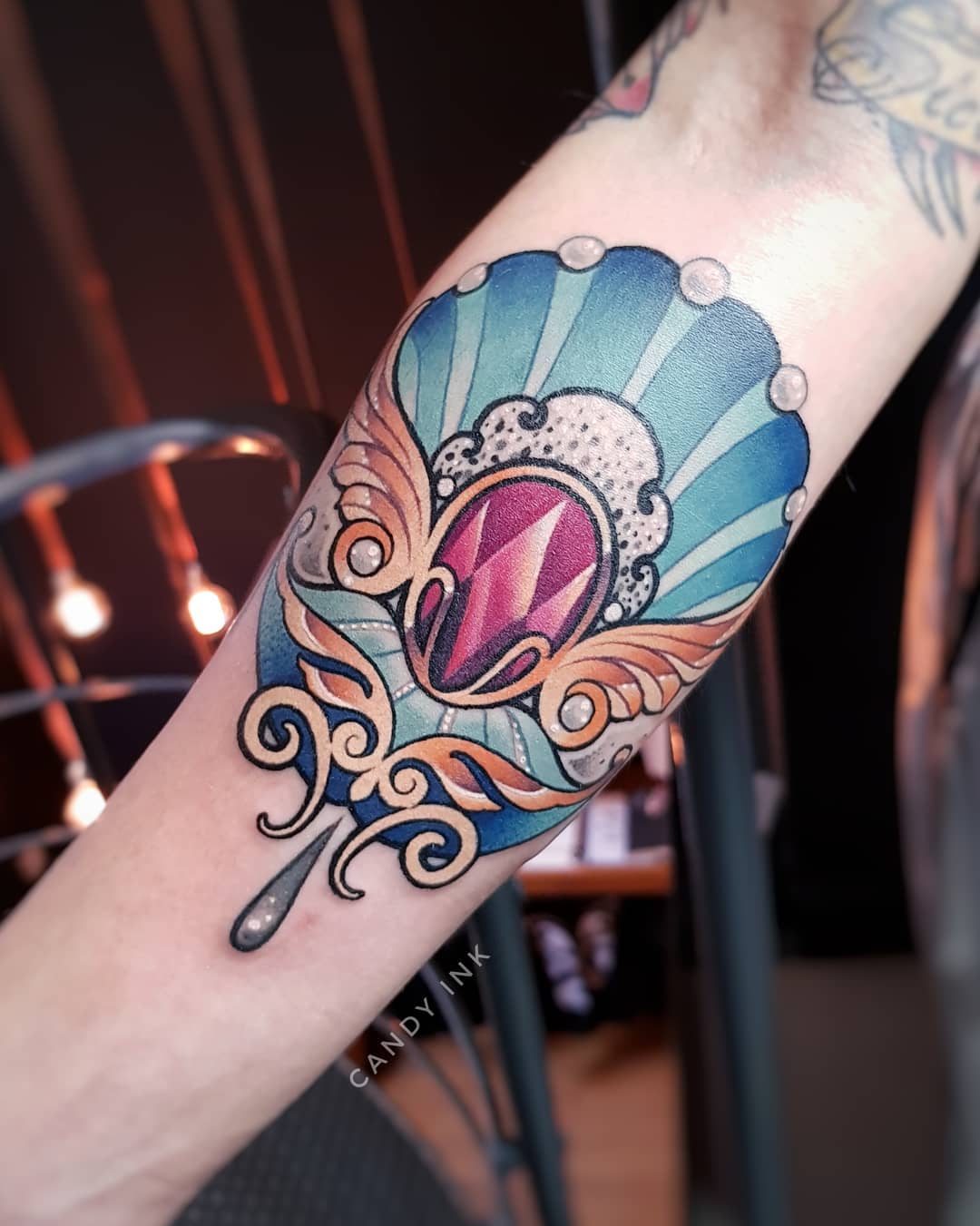 a neo traditional style tattoo of a jewel on a forearm. the jewel is pink and purple and is surrounded by gold ornate wings and a turquoise seashelllike backing. the tattoo is detailed and colorful.
