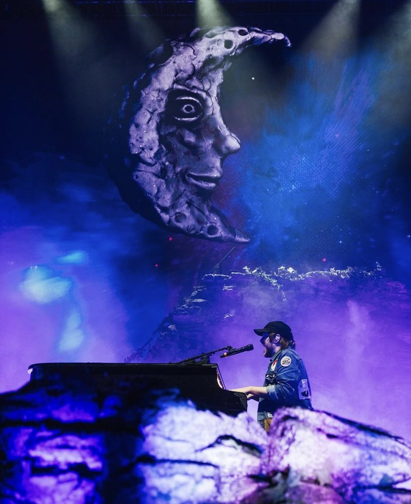 a musician is performing on stage with a piano in front of a giant moon prop. the stage is lit in purple hues and the musician has a microphone in front of him.