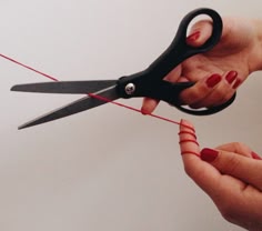 a closeup shot of a hand holding a pair of scissors, cutting a red thread with a finger wrapped in it. the hand is decorated with red nail polish.