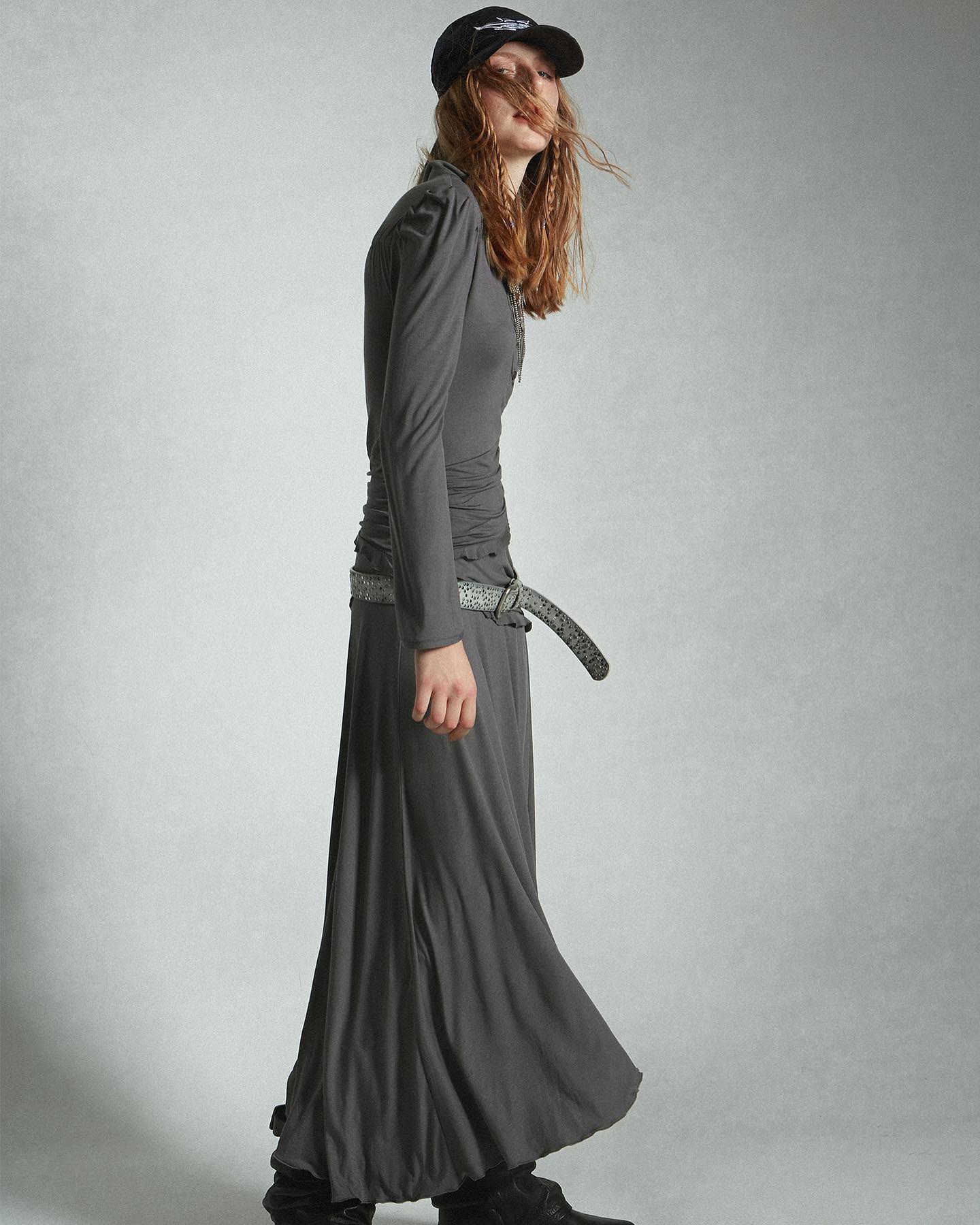 a fulllength image of a female model showcasing a stylish grey longsleeved dress with a long, flowing skirt. the dress is cinched at the waist with a thin belt and paired with dark shoes and a cap. the model is posed in a side profile against a light grey backdrop.  the overall aesthetic is contemporary and elegant.