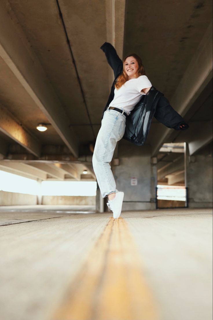 a young woman jumps in the air, smiling and laughing, while wearing a white tshirt, blue jeans and white sneakers. she's wearing a black jacket over her shoulders. the background is a concrete underpass with yellow lines on the ground. she is jumping towards the camera.