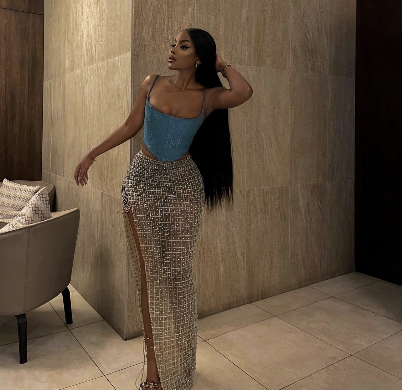 a beautiful black woman is posing for a fashion shoot. she is wearing a blue corset top and a glamorous beaded skirt. her long, black hair cascades down her back and she is standing against a wall. the image is full of glamor and style, with a focus on the woman's elegant pose and her beautiful clothing.
