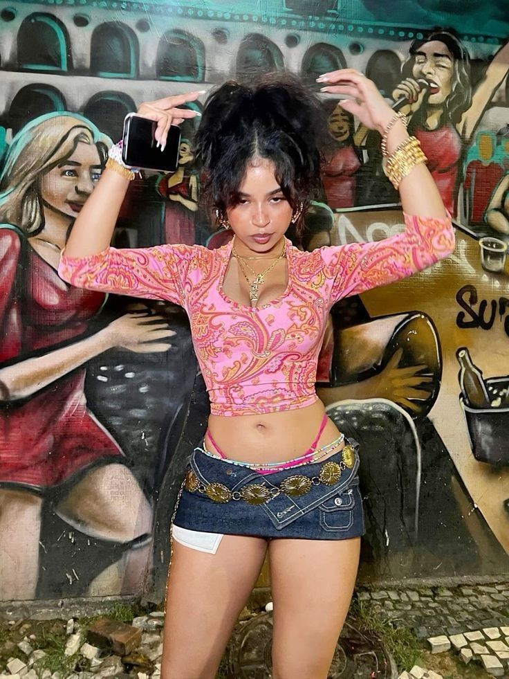the image features a young woman confidently posing in front of a vibrant graffiti mural. she's wearing a pink paisley longsleeve crop top, a denim mini skirt, and a large, ornate belt. her accessories include gold jewelry and a high ponytail hairstyle. she holds a cell phone in one hand and is styled in what appears to be a 90sinspired or y2kinspired fashion style. the background mural depicts a lively scene with several women. the overall mood is one of youthful energy and selfassuredness.