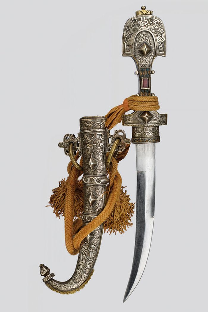 an ornate middle eastern jambiya dagger is displayed with its silver scabbard and hilt. the dagger has a curved blade and the scabbard is intricately decorated with silver designs. a thick, goldenbrown cord with tassels wraps around the dagger and scabbard, adding to its elaborate appearance. the background is a plain, light color.