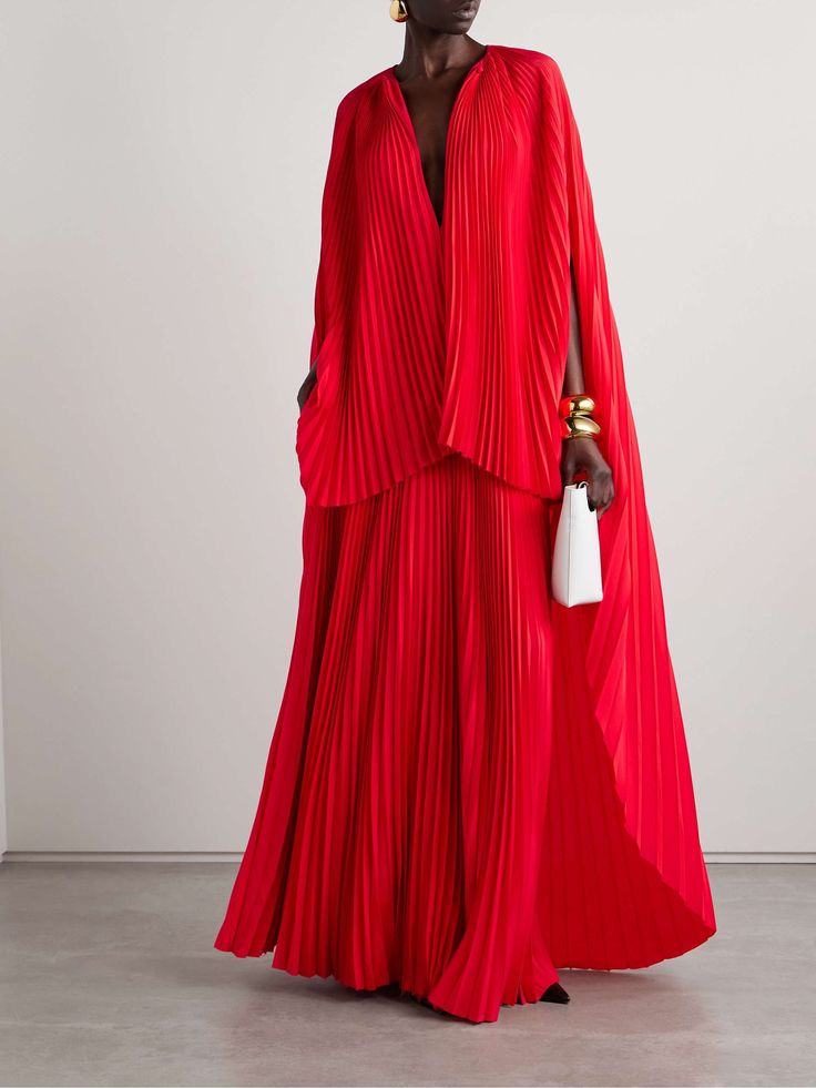 a woman wears a long red pleated cape dress, posing for a photo shoot against a white background.  she wears gold earrings and a few gold bracelets on her right wrist, and holds a white handbag with a gold clasp.  she has her hair pulled back, revealing a nude makeup look.