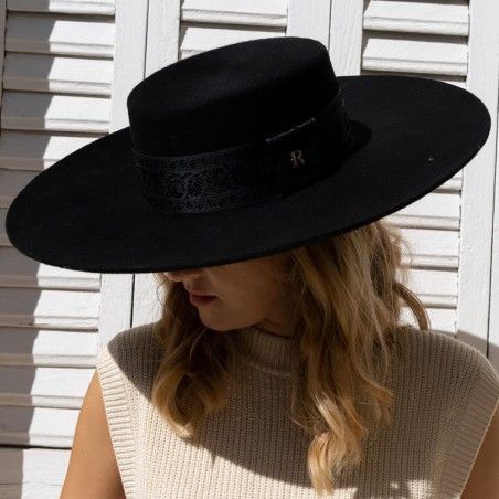 a woman in a black widebrimmed hat with a black band, posing with the hat covering her eyes, looking down, she is wearing a beige sweater and the wall behind her is made of white painted wood slats.  the woman is looking down and the light is casting shadows on her face.