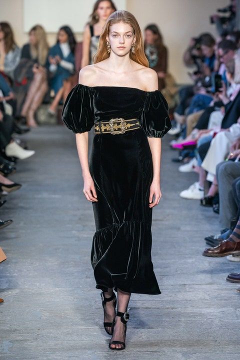 a model walks the runway wearing a black velvet offtheshoulder midi dress with puff sleeves. the dress has a ruffled hem and is accessorized with an ornate gold belt. she is wearing black strappy heels. the background is blurred, showing other models and audience members. the overall atmosphere is dark and elegant.