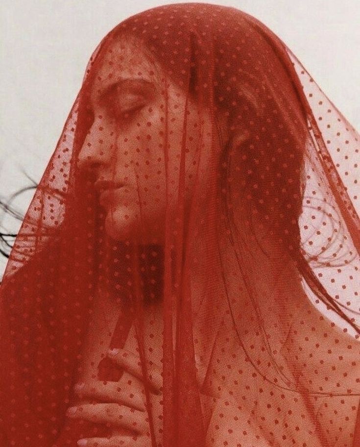 a woman with long dark hair is wearing a red mesh veil that covers her face and hair. the veil is sheer and allows glimpses of her features, creating a sense of