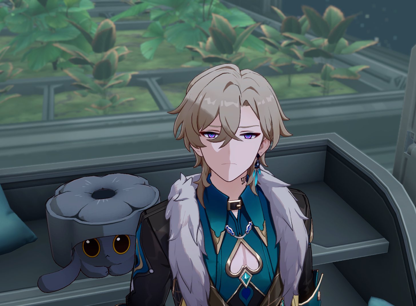 a closeup shot of scaramouche from genshin impact.  he is a young man with blond hair, purple eyes, and is wearing a teal and dark colored outfit with a white fur collar. he has an earring and necklace, and a small tattoo is visible on his neck.  he appears to have a serious or unhappy expression. in the foreground is a small gray plush toy that resembles a cat wearing a flowershaped hat.  the background shows an indoor setting with shelves and potted plants.