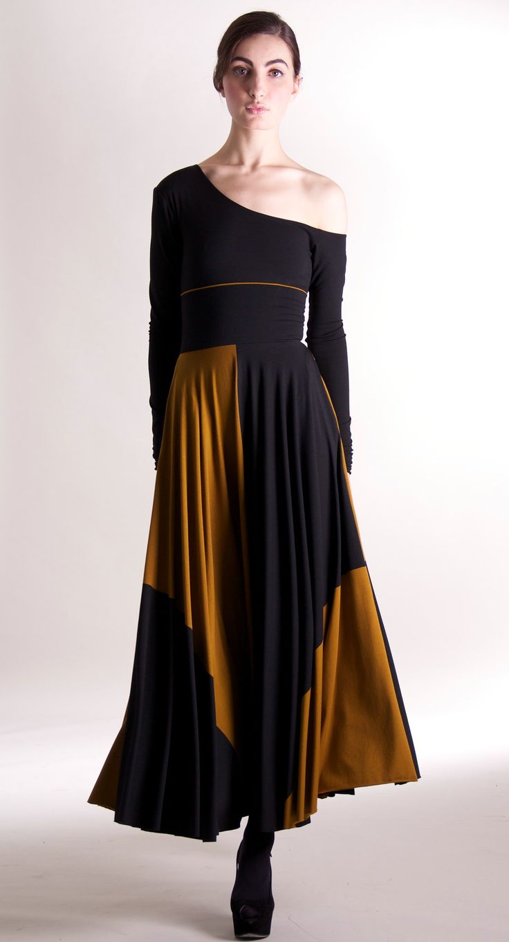 a woman in a black and yellow dress is standing in a studio, looking at the camera. the dress is long and has long sleeves. she is wearing black heels. the dress is a oneshoulder style with a black bodice and a gold skirt. she is a fashion model.