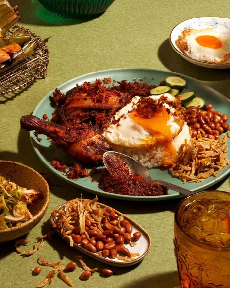 a closeup overhead shot of a plate of nasi lemak, a popular malaysian dish. the vibrant image shows the dish's key components: fragrant rice, crispy fried chicken, a perfectly fried sunnysideup egg, crunchy peanuts, and sliced cucumbers.  a small bowl of sambal (chili paste) is visible, along with other side dishes such as fried anchovies and more peanuts.  other food items like a bowl of what appears to be a salad and a  basket of banana leaves with something inside are in the background, suggesting a full meal.  the setting is casual yet elegant with a green tablecloth and other bowls and a drink adding to the scene. the overall composition and lighting creates a mouthwatering and appealing visual representation of the meal.