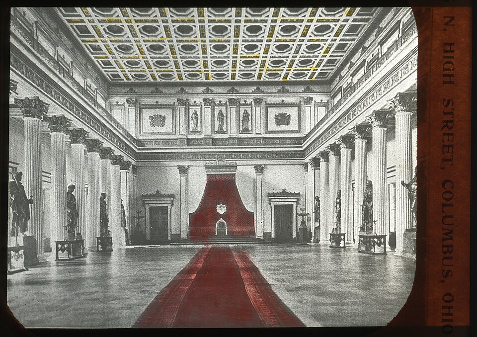 a historic photograph shows the interior of a grand building with a red carpet leading to a raised platform with a red backdrop. the hall is lined with columns and statues. the ceiling has an ornate pattern. text along the right side reads "n. high street, columbus, ohio". it appears to be the ohio statehouse.