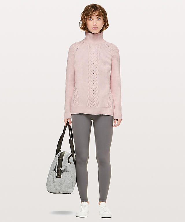 a woman is standing in front of a white background, modeling a pink cable knit turtleneck sweater, grey leggings, and white sneakers. she is also carrying a grey gym bag with black accents on her left shoulder.