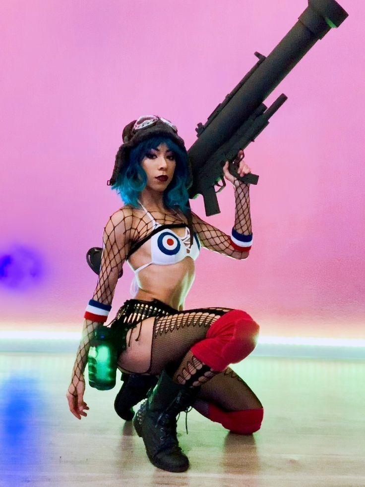 a woman in a cosplay costume is kneeling with a rocket launcher in her hand. she is wearing black fishnets and red and blue bracelets on her wrists. she is looking at the camera with a serious expression on her face. she is in a studio with a pink background.