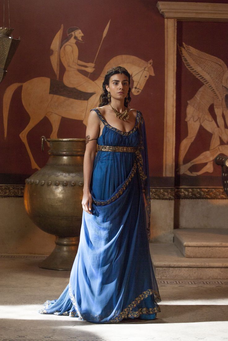 a woman in a flowing blue ancient roman or greek style gown stands in a room with painted murals on the wall.  she wears gold jewelry and her expression is serious.  the setting appears to be a grand room or palace.  a large bronze urn is partially visible in the foreground.