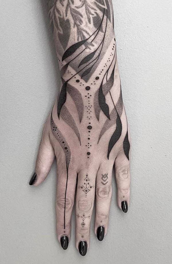 a hand and lower arm are shown with an elaborate black ink tattoo design. the design features a combination of geometric shapes, dotwork shading, stylized leaves, and ornamental elements, covering much of the hand and lower arm. the tattoos extend onto the fingers, with smaller decorative patterns. the overall aesthetic is intricate and artistic.