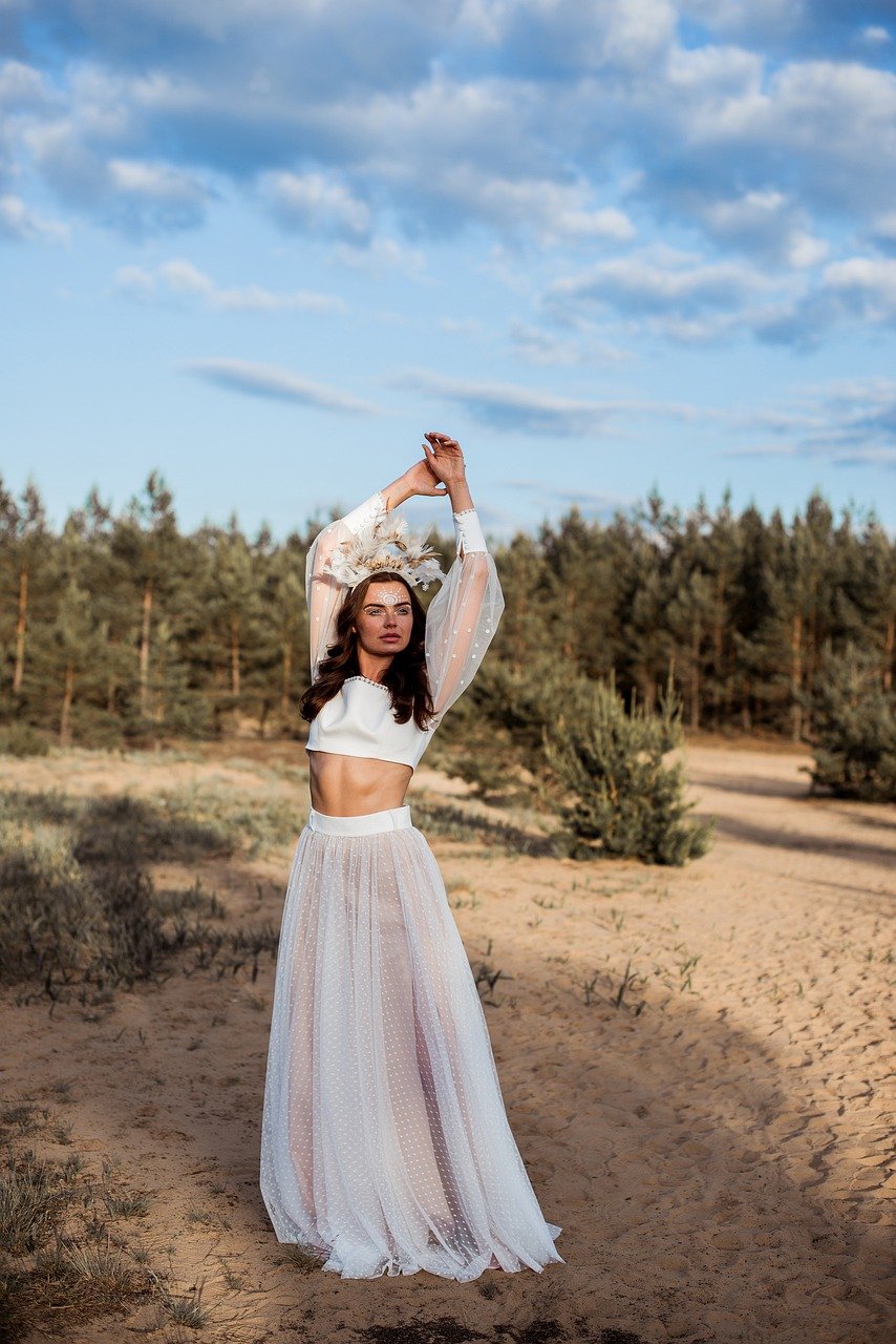 a fulllength shot of a woman model wearing a beautiful white bohostyle outfit.  the outfit consists of a fitted white crop top and a long, sheer tulle skirt with a subtle polka dot pattern. she also wears a delicate floral headpiece. her arms are raised above her head. she is posing in a sandy, desertlike landscape with a forest in the background under a partly cloudy sky. the overall mood is elegant, stylish and serene.