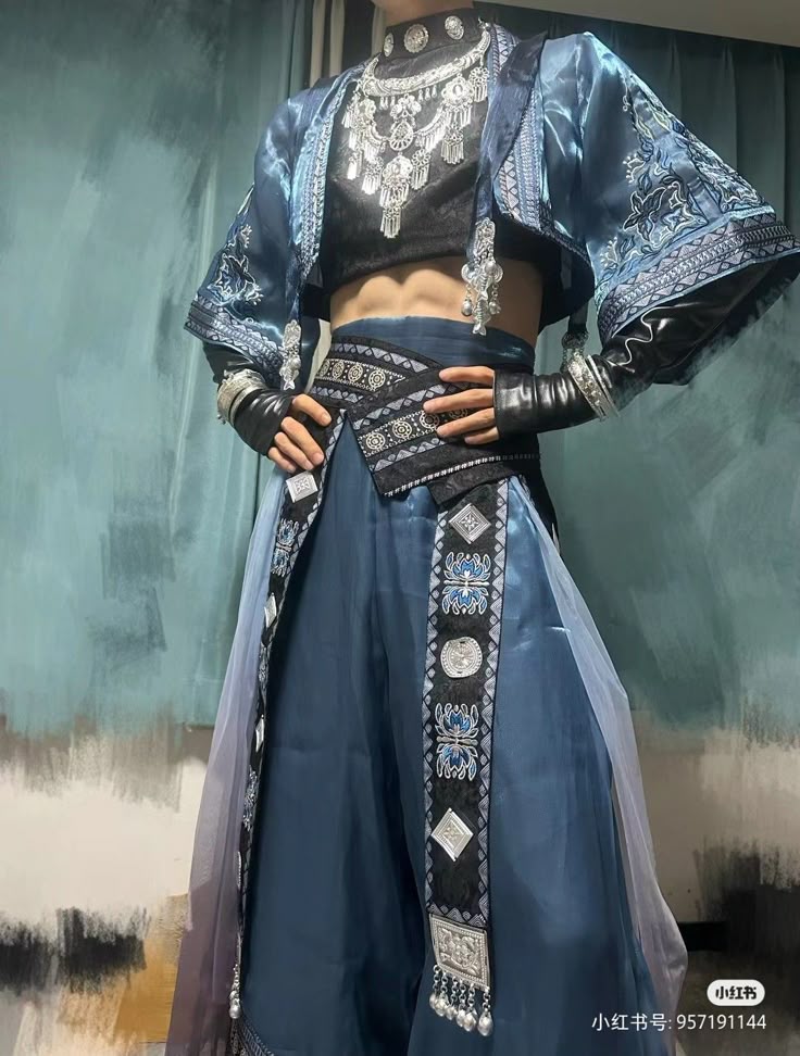 a person is posing wearing a unique and ornate clothing set. the outfit includes a black crop top, a blue skirt with a decorative sash, and a blue jacket with embroidered details. they are also wearing multiple pieces of jewelry and black leather arm coverings. their hands are resting on their hips, and their abs are visible. the background is a mottled blue.