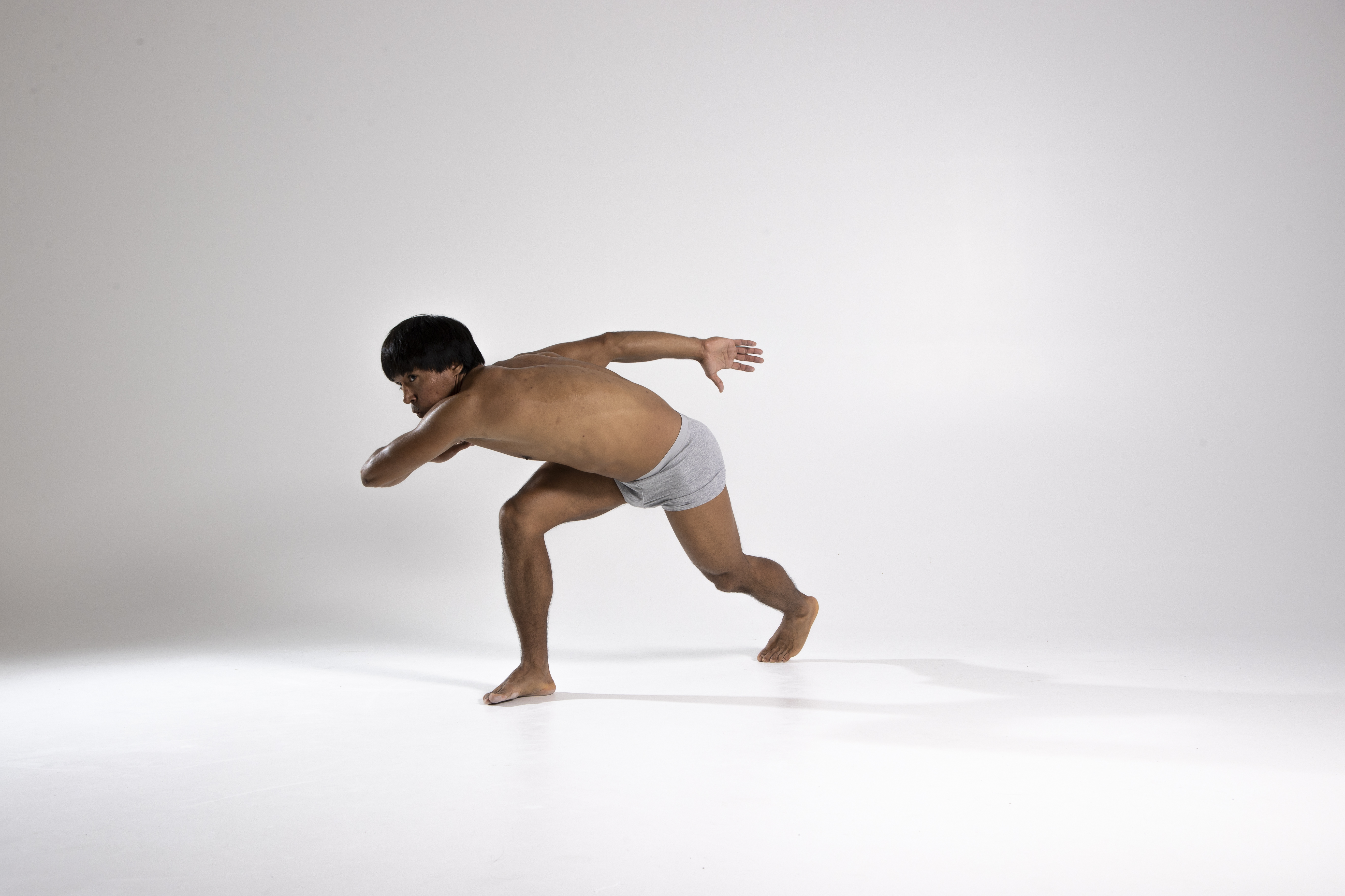 a shirtless man wearing grey underwear is captured in a studio shot against a plain white background. he is barefoot and in motion, leaning forward with his arms outstretched, creating a dynamic and athletic pose. his dark hair contrasts with his tan skin, adding to the visual impact of the image.