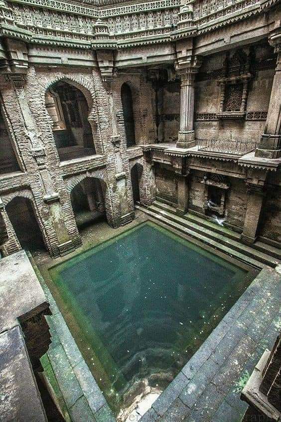 a highangle view of an ancient stepwell, showcasing its intricate architecture, including arches, pillars, and ornate stone carvings. the stepwell has a rectangular pool of water at the bottom, and steps leading down to the water level. the structure appears to be made of stone and reflects a historical site, potentially located in india.