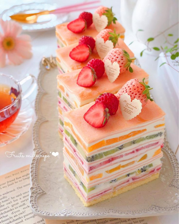 the image shows slices of fruit mille crepe cake on a silver serving tray. the mille crepe cake consists of multiple layers of crepes and cream, with various types of fruit such as strawberries, raspberries, and kiwi layered in between. each slice is topped with fresh strawberries, raspberries, and a small paper tag that reads "handmade sweets". a cup of tea is also visible on the side of the tray, along with a plate and utensils in the background. the composition is bright and airy, with a soft focus. the scene is set on a table with a book underneath, with some of the text on the book showing and appearing to be from a harry potter book. the cake is labeled "fruits millecrepe" in the bottom left corner.