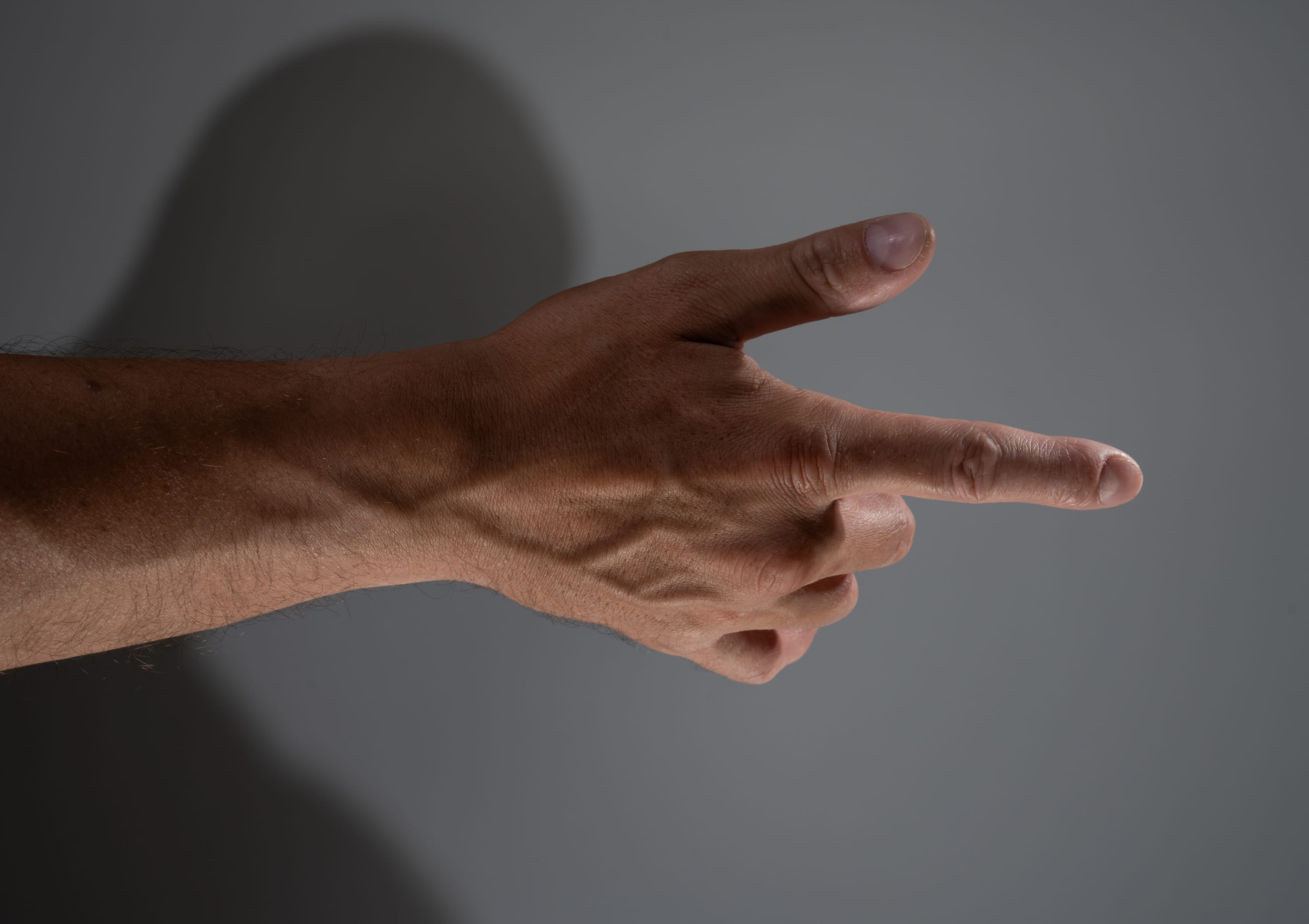 a man's arm and hand extended in front of a grey background, with his index finger pointing towards the right. the hand is illuminated by a studio light, highlighting the details of the skin and veins.