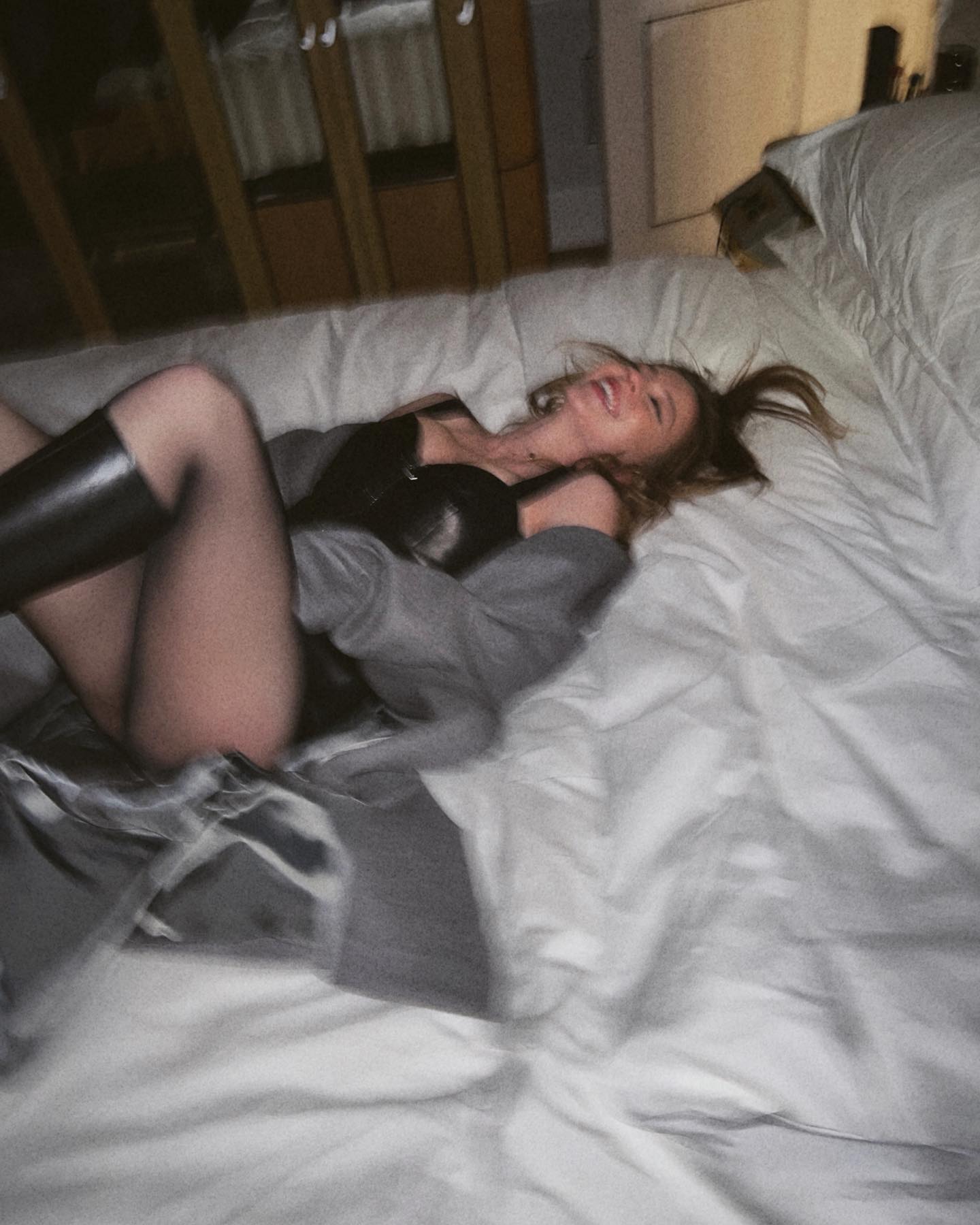 a blurry, intimate photograph of a young woman lying playfully on a bed. she's wearing black lingerie, a grey cardigan, and black boots. her hair is tousled, and she's smiling and laughing. the setting is a dimly lit bedroom, and the overall mood is sensual and relaxed. the image has a slightly grainy, filmlike quality, contributing to its moody and intimate atmosphere.