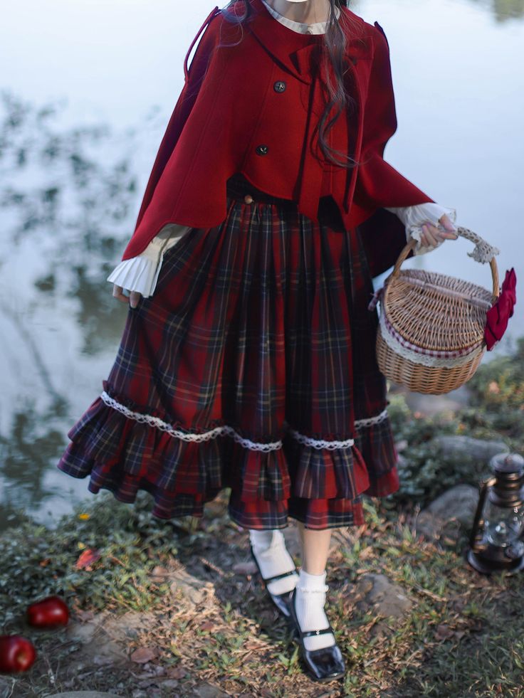 a woman wearing a red cape, white blouse with lace cuffs, and a red and navy plaid skirt stands near a lake. she holds a wicker basket with a red flower in it. the image evokes a fairytale, red riding hood aesthetic. the environment suggests a forest setting, with fallen apples and a lantern on the ground nearby.