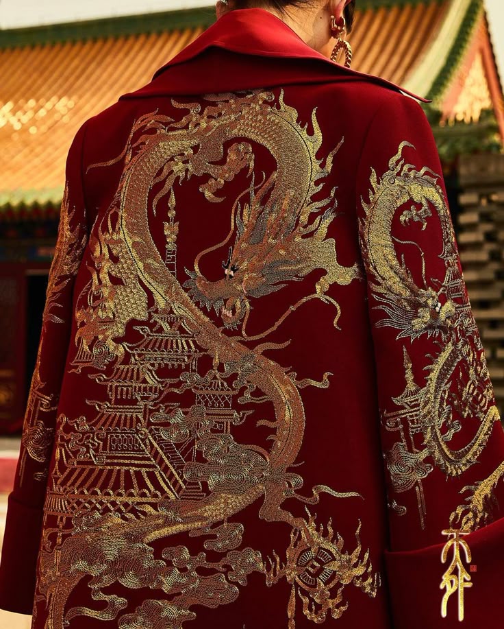 a closeup shot shows the back of a red coat with an elaborate golden dragon embroidery. the dragon winds around a detailed depiction of a traditional chinese building, possibly a temple or palace. the embroidery features intricate patterns and accents, suggesting a luxurious and culturally significant garment. the collar and sleeve details of the coat are visible, and the background shows glimpses of asian architecture.