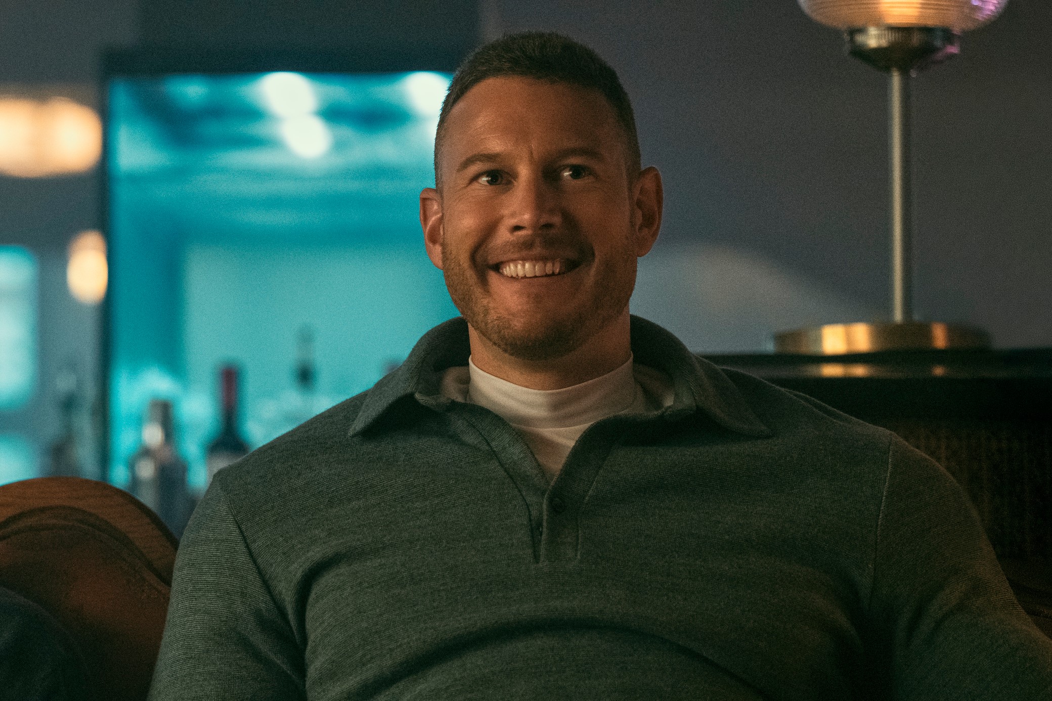 a man with short hair and a slight smile, wearing a green collared shirt, is sitting on a brown leather couch in a dimly lit room. he is looking directly at the camera.  a bar cart with bottles on it is behind him to the left and a lamp with a round shade is behind him to the right.