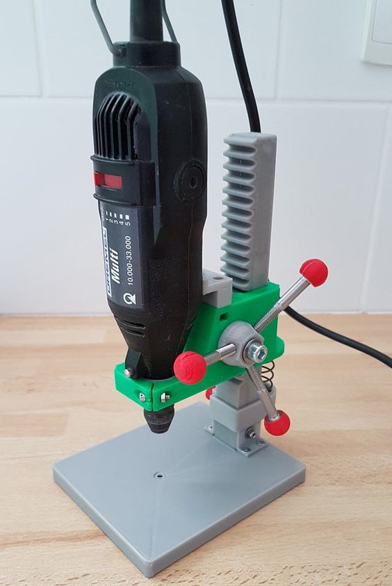 a 3d printed drill press holding a rotary tool. the drill press is made of grey plastic, with a green base. the tool is black and has a red power switch. the drill press is sitting on a wooden surface.