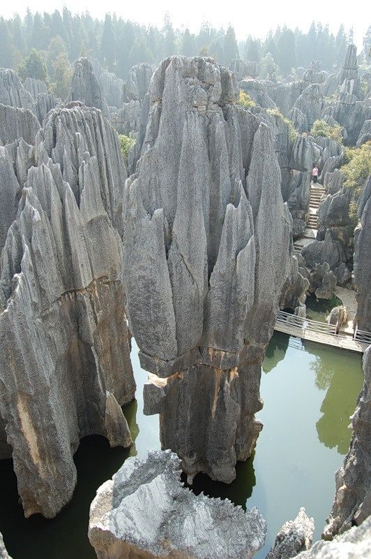 a scenic view of the stone forest in yunnan, china, showcasing towering karst rock formations reflected in a lake. a wooden walkway and stairs wind through the landscape, offering a path for visitors to explore the unique natural formations. the background includes trees and a hazy sky, adding to the overall landscape.